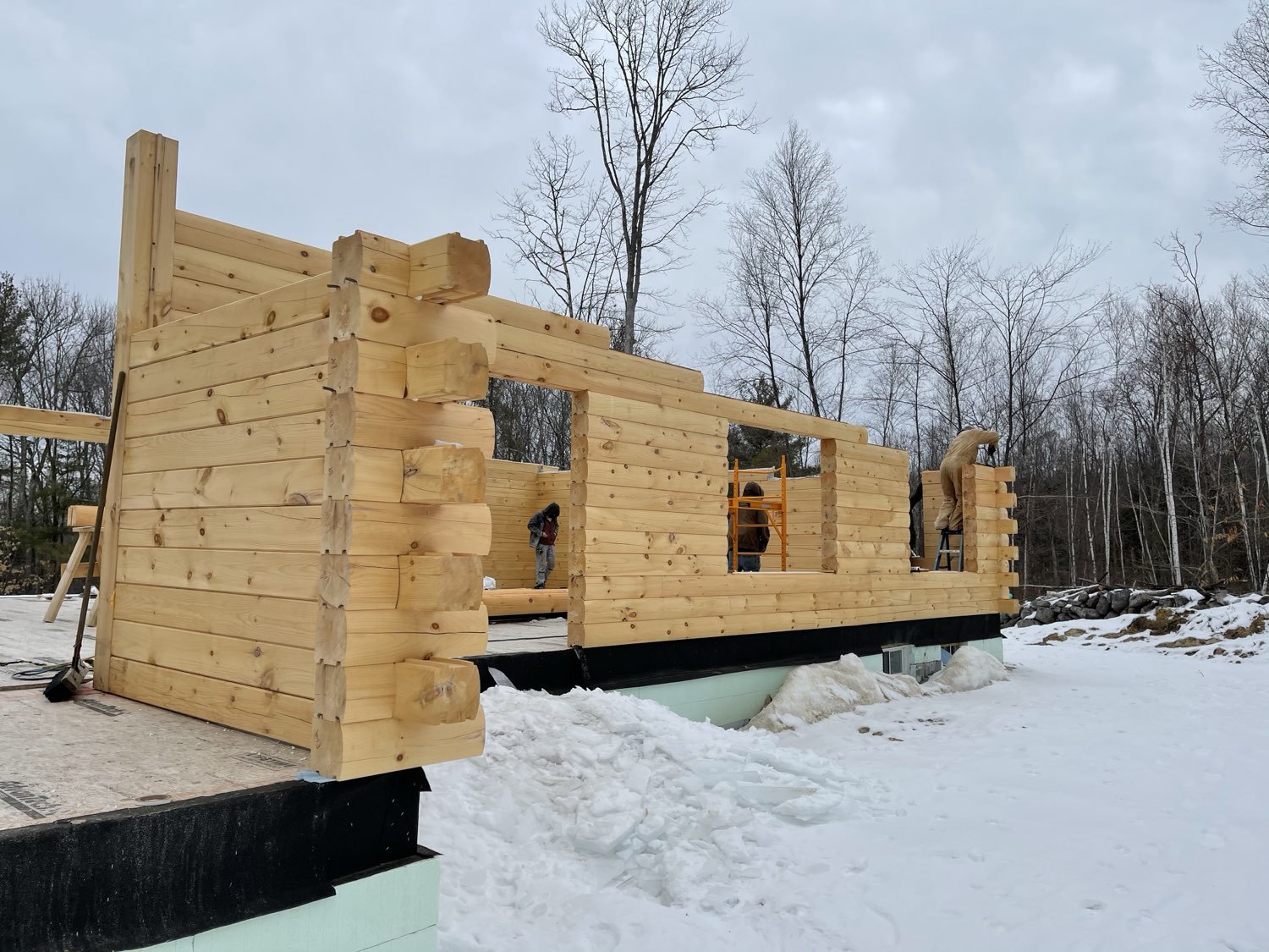 A Merrimac log home package being assembled on site in New Hampshire, with the log walls up and a crew finishing the chimney and roof