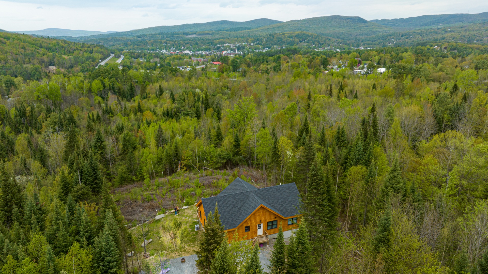 Aerial view of a finished Sunapee log home in a forest clearing with mountain valley and town beyond
