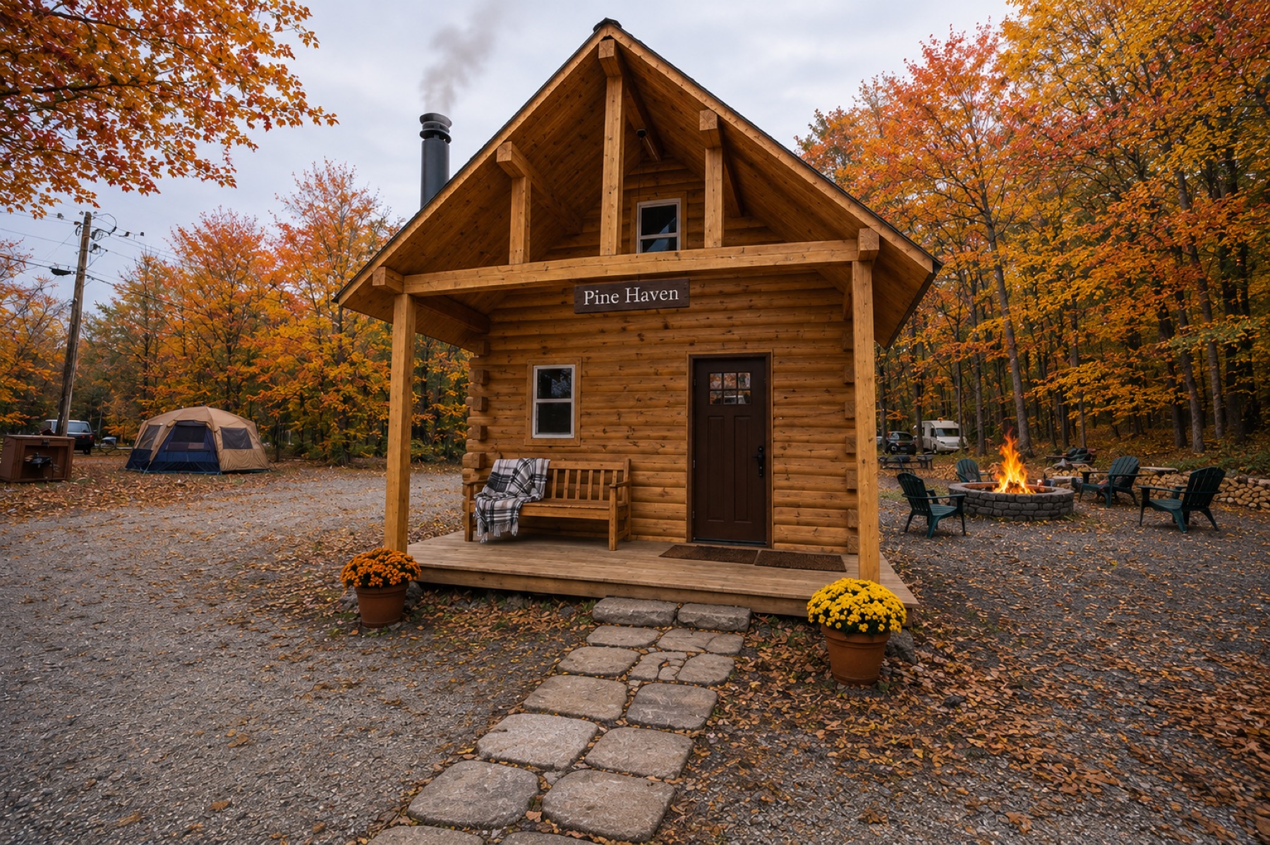 The Pine Haven tiny log cabin in an autumn campground with a tent, a stone fire ring, and Adirondack chairs in the background